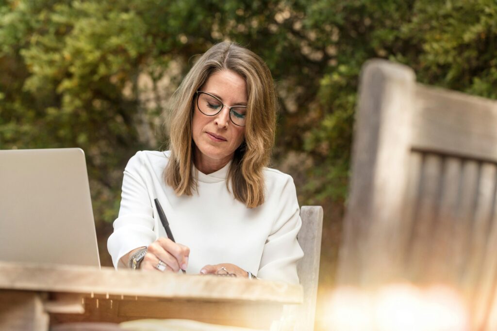 A woman in glasses writing and working on her laptop outdoors in natural daylight.
